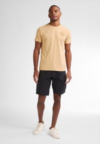 Tan katoenen T-shirt met een ronde hals en een grafische print aan de voorkant, gecombineerd met zwarte cargoshorts met zijzakken. Afgemaakt met witte sneakers.