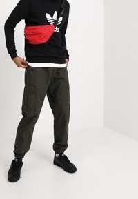 Svart sweatshirt med vit Adidas-logga, röd midjeväska, olivgröna cargobyxor och svarta sneakers med vita detaljer. Enkel bakgrund.