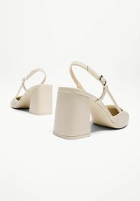 Sandales à talons beiges avec un design à T, en matériau synthétique lisse, talon large et géométrique, et bride de cheville ajustable.