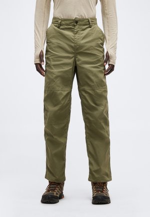 Person trägt olivgrüne Cargohose, beiges langärmeliges Hemd und braune Wanderschuhe, steht vor einem einfarbigen hellen Hintergrund.