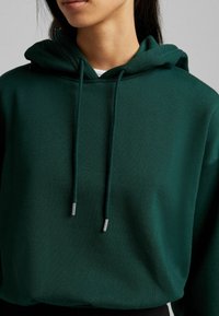 Sudadera verde con una textura suave, que presenta un cordón con puntas metálicas. El diseño incluye un corte relajado y una capucha.