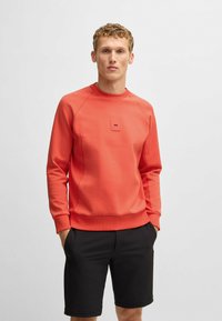 Korallefarbener Pullover mit Rundhalsausschnitt und gerippten Bündchen, mit einem kleinen quadratischen Logo auf der Brust. Getragen mit schwarzen Shorts.