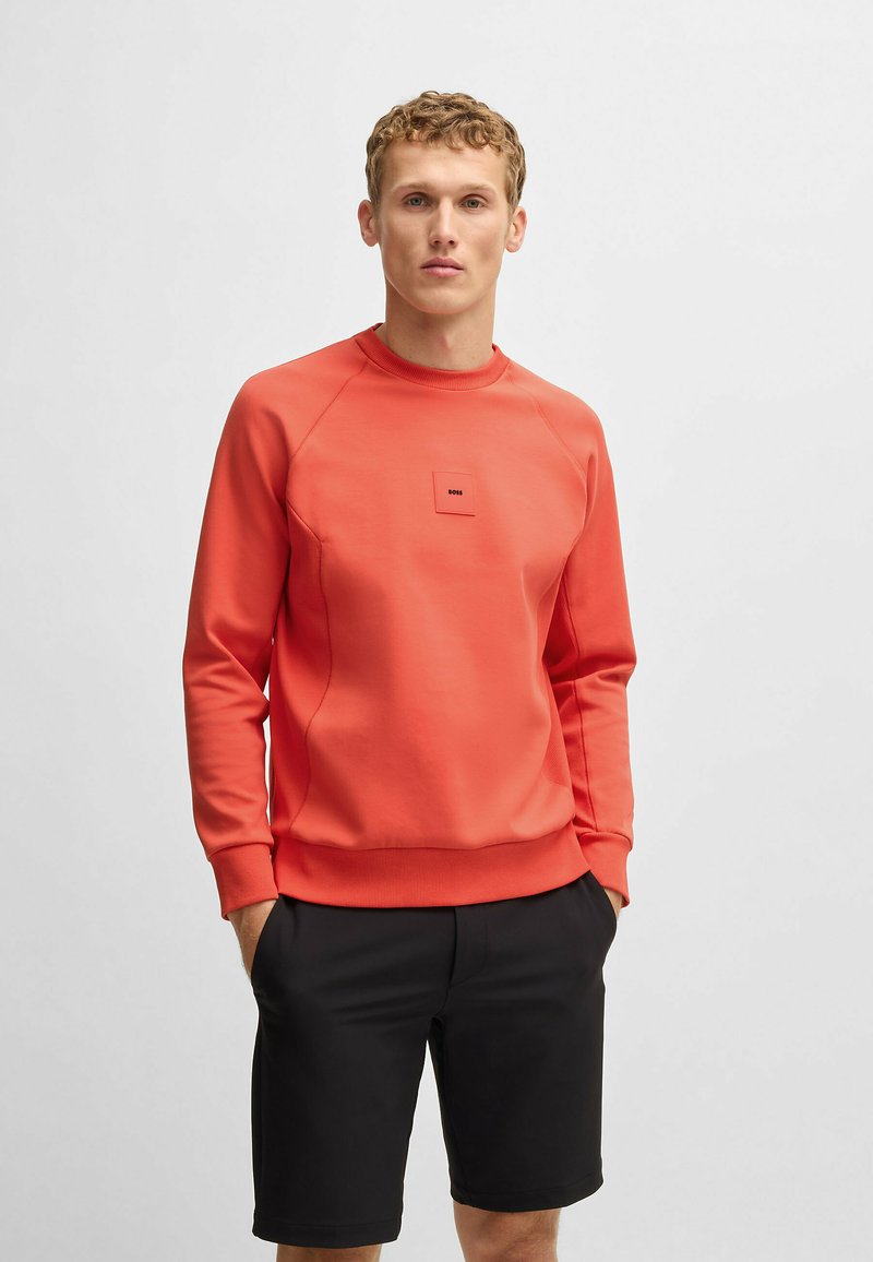 Korallefarbener Pullover mit Rundhalsausschnitt und gerippten Bündchen, mit einem kleinen quadratischen Logo auf der Brust. Getragen mit schwarzen Shorts.