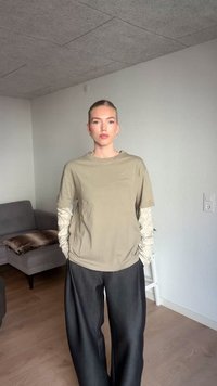 Mlada žena stoji unutra, odjevena u bež slojevitu dugosvjetlu majicu i široke crne hlače, s rukama u džepovima, neutralnog izraza lica.
