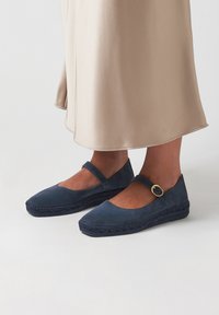 Tmavě modré espadrilky s plochým designem, zaoblenou špičkou, páskem se zlatou sponou a tkanými juty akcenty na podrážce.
