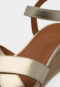 Χρυσαφένιο μεταλλικό σανδάλι πλατφόρμα με λουριά που διασταυρώνονται και καφέ επενδεδυμένη ανατομική σόλα.