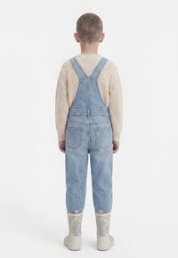 Otrok obrnjeno stran, oblečen v svetlo modre jeans nepredušne hlače na naramnice, bež pleten pulover in škornje s cvetličnim vzorcem, stoji na belem ozadju.