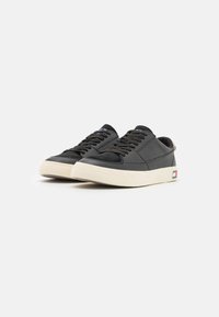 Zapatillas negras de cuero texturizado, con cordones planos y suela de goma color crema; presentan un pequeño parche de logo en rojo, blanco y azul en el lateral.