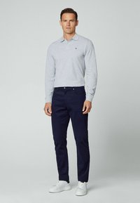 Graues Langarm-Polo mit Kragen, marineblaue Hose und weiße Sneaker. Das Polo hat ein kleines Logo auf der Brust.