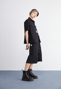 Black short-sleeve shirt over a light blue top, paired with black cargo shorts and chunky black lace-up boots. The look is accessorized with a striped bucket hat.