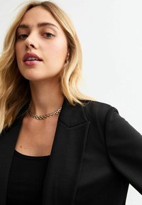 Blazer negro con hombros estructurados y una tela texturizada. Combinado con una blusa negra y un collar de cadena dorada, con un acabado suave.