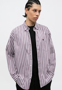 Fiatal férfi fonott hajjal, egy nagy méretű, lila és fehér csíkos inget visel egy fekete póló felett, kezét a zsebébe dugva, oldalra néz.