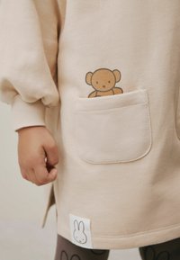 Beżowa bluza z bufkami i kieszenią, z ilustracją brązowego misia. Udekorowana białą metką przedstawiającą twarz królika.