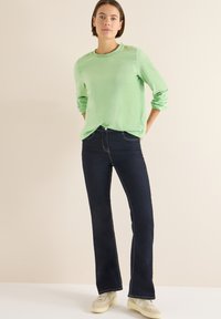 Zielona bluzka z długim rękawem o fakturowanej tkaninie, z okrągłym dekoltem, noszona z ciemnoniebieskimi dzwonowatymi jeansami i jasnymi trampkami.