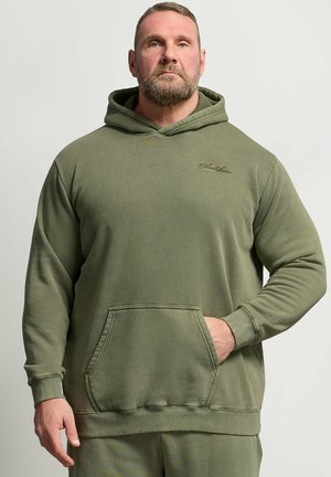 Bearded man wearing olive green hoodie with front pocket, standing against a plain light background, hand partially in pocket.