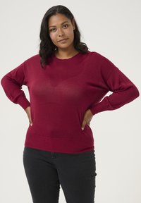 Pull en tricot rouge avec un col rond, coupe décontractée et poignets côtelés. Le matériau a un léger éclat, associé à un jean sombre.