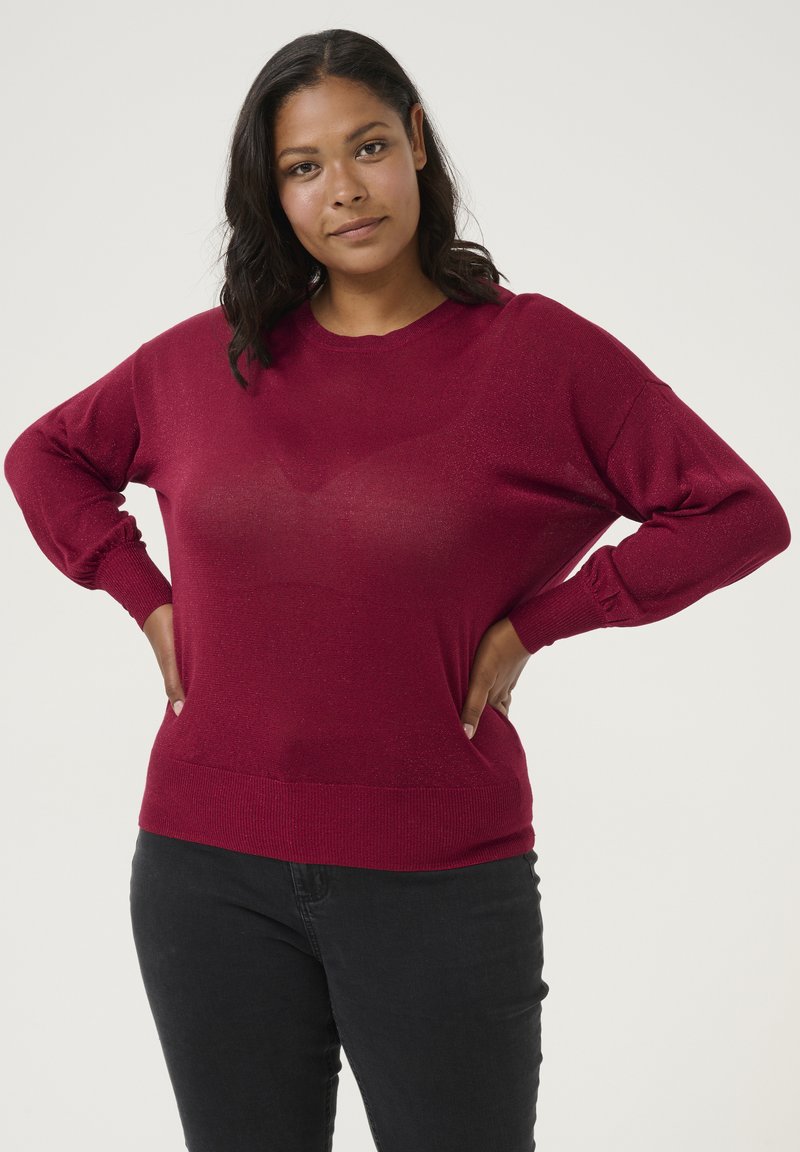 Pull en tricot rouge avec un col rond, coupe décontractée et poignets côtelés. Le matériau a un léger éclat, associé à un jean sombre.