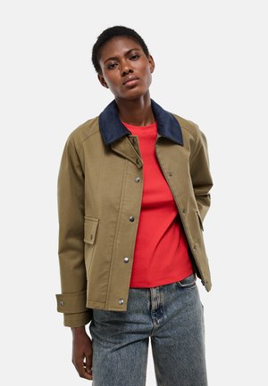 Jeune femme noire portant une veste olive avec un col bleu marine sur une chemise rouge et un jean bleu, debout devant un fond clair uni.