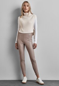 Crème ribgebreide trui zonder mouwen gecombineerd met strakke beige faux leren leggings en witte sneakers met lichtbruine zolen.
