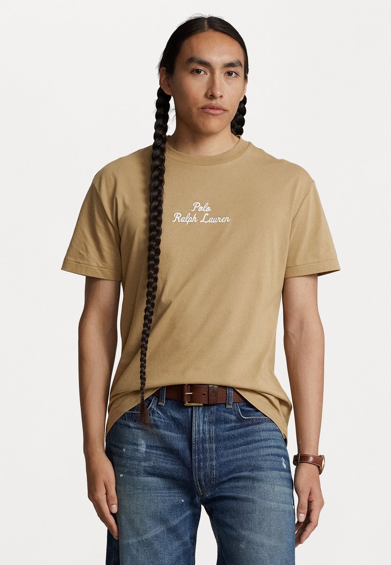 Hnedá bavlnená tričko s bielym logom "Polo Ralph Lauren", v kombinácii s modrými džínsami a hnedým opaskom. Model má dlhé, tmavé vrkoče.