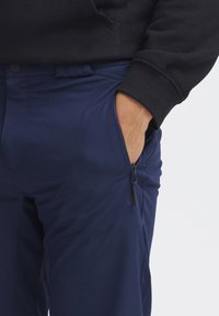 Námořnické modré kalhoty s boční kapsou na zip, hladkým materiálem s mírným leskem a strukturovaným páskem. V kombinaci s černou mikinou.