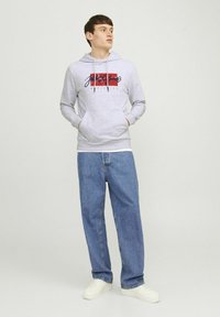 Grijze hoodie met een voorvak, met een print van een rood en marineblauw logo. Combinatie met losse blauwe denim jeans en witte sneakers.