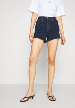 Femme portant un short en jean bleu foncé effiloché, un T-shirt blanc, des sandales noires à talons et un petit collier pendentif rond, debout sur un sol gris.