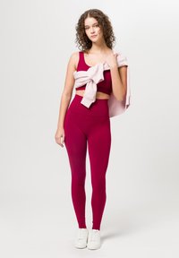 Bordeaux cropped top en high-waisted leggings, gecombineerd met een lichtroze sweatshirt dat over de schouders is gedrapeerd en witte sneakers.