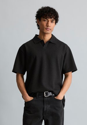 Junger Mann mit lockigem dunklem Haar, der ein schwarzes, strukturiertes Kurzarm-Poloshirt und schwarze Jeans trägt, die Hände in den Hosentaschen.