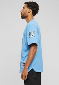 Šviesiai mėlyna trumpomis rankovėmis marškinėliai su apvalia iškirpte, su grafiniu dizainu priekyje ir dekoratyvine aplikacija ant kairės rankovės.