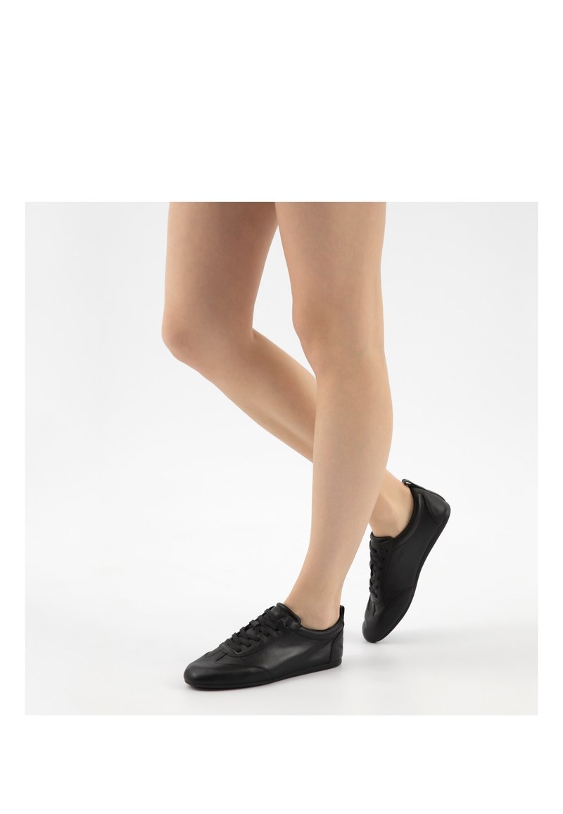 Beine in schwarzen, flachen Leder-Sneakers, die auf einer weißen Oberfläche stehen, wobei ein Fuß leicht hinter dem anderen angehoben ist.