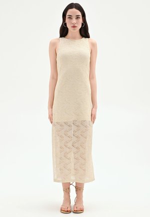 Femme portant une robe midi en maille beige sans manches avec un motif en zigzag et une partie inférieure transparente, debout les bras le long du corps sur fond blanc.