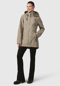 Bežs āra jaka ar kapuci, ar auklām, ar rāvējslēdzēja aizdari, divām priekšējām kabatām un teksturētu apdari. Valkāta virs melnajām biksēm.