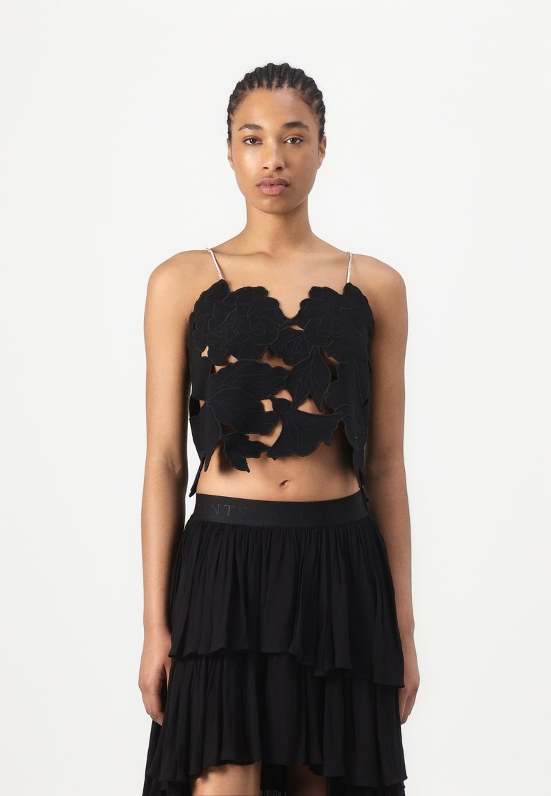 Frau mit geflochtenem Haar trägt ein schwarzes florales Crop-Top mit dünnen Trägern und einen gestuften schwarzen Rock vor einem schlichten weißen Hintergrund.