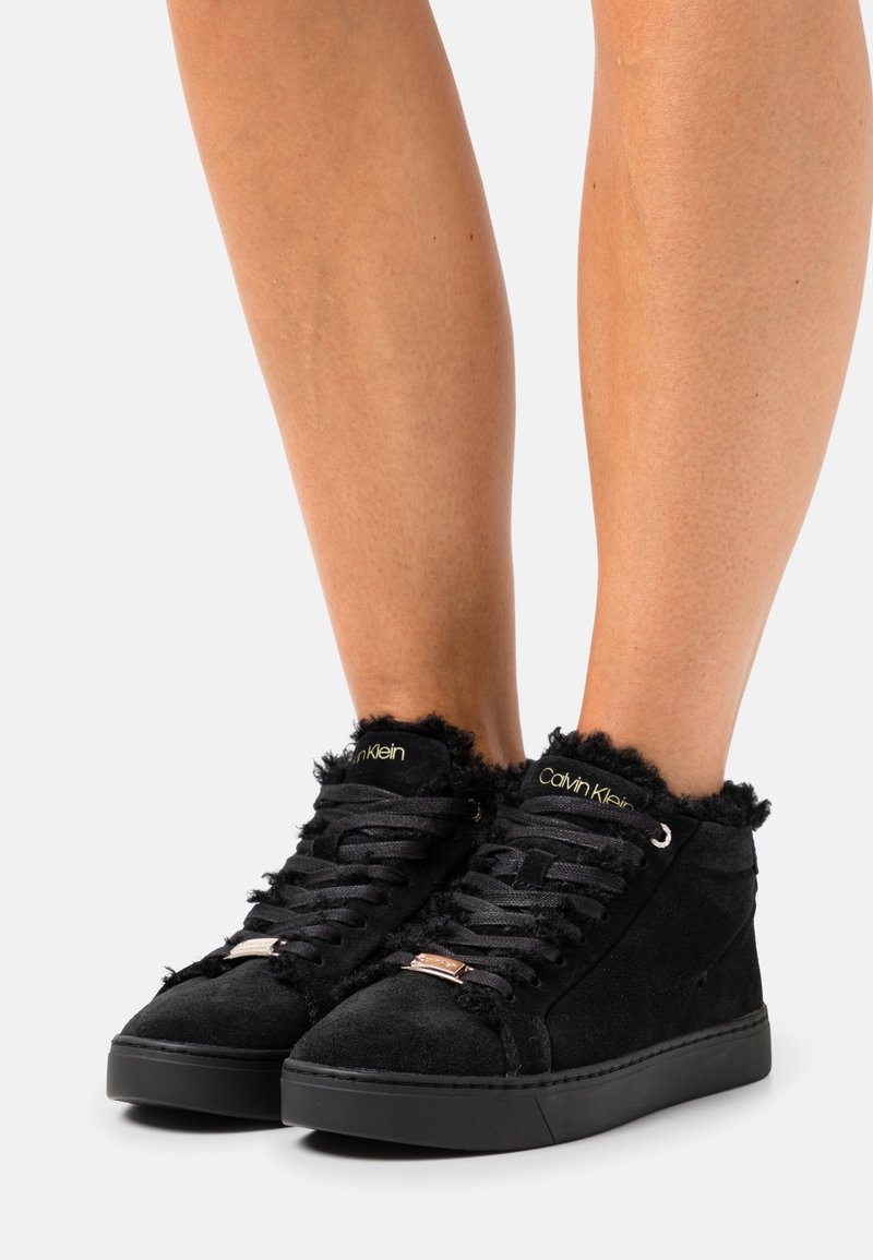 Baskets montantes en daim noir avec lacets noirs et bordure en fourrure portées sur des jambes nues sur fond blanc.