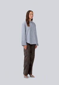 Camisa de botões às riscas em azul e branco, combinada com calças largas de cor castanha escura. Apresenta punhos largos e um único bolso no peito.