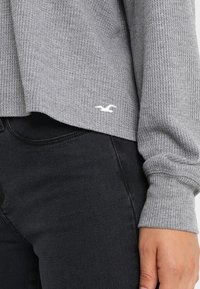 Harmaa ribbed-neule cropped-mallisilla pitkähihaisilla, jossa on valkoinen brodeerattu logo vasemmassa alareunassa. Yhdistetty mustiin farkkuihin.