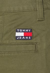 Tessuto verde oliva con una toppa cucita che presenta "TOMMY JEANS" in bianco e blu, con strisce rosse e bianche, situata su una tasca.