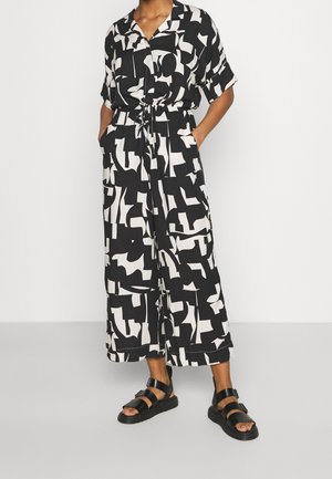 Combinaison noire et blanche à motifs abstraits, manches courtes, boutonnée sur le devant, taille ajustable par cordon, avec poches latérales. Portée avec des sandales plates noires.