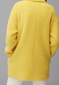 Abrigo amarillo suave con un corte relajado, mangas largas y un cuello ancho. La textura parece suave y cálida, sin herrajes visibles.