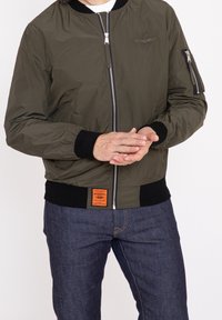 Olivgrön bomberjacka med dragkedja fram, ribbad muddar och midja. Har sidofickor och en liten logotyp på bröstet.