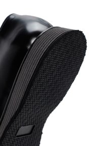 Semelle de chaussure en cuir noir avec une texture en caoutchouc antidérapant et un bord courbé. Présente un motif en croix subtil sur le côté.