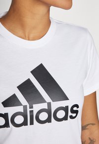 Hvit bomull T-skjorte med et stort svart Adidas-logo. Kragen er rund og ribbet, med korte ermer. Bitt av tekstur er observert.