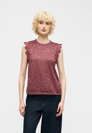 Junge Person mit lockigem blondem Haar, trägt ein ärmelloses lila Spitzenoberteil und dunkelblaue Hose, steht vor einfachem Hintergrund.