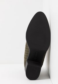 Semelle de chaussure noire à talon avec grip texturé, inclinée contre un mur blanc d’angle, affichant la taille 37 et la marque.