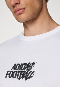 Hvid bomulds t-shirt med et sort grafisk tryk af "ADIDAS FOOTBALL" over brystet, med rund halsudskæring og korte ærmer.