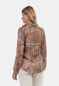 Blusa a maniche lunghe in tessuto stampato, caratterizzata da intricati motivi paisley e geometrici nei toni del marrone, arancione, blu e bianco.