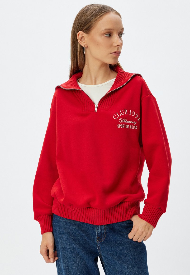 Czerwony sweter z półzamkiem, z prążkowanymi mankietami i dołem, z haftem "CLUB 1994 Williamsburg SPORTING GOODS" w kolorze białym.