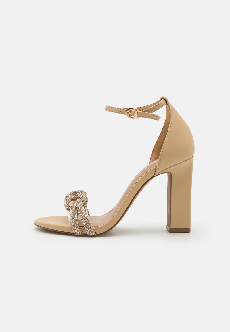 Beige høyhælte sandaler med firkantet tå, tykk blokkhæler og en dekorativ knute med metalliske aksenter på frontstroppen.