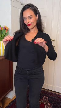 Femme aux longs cheveux foncés, vêtue d'un haut noir et d'un jean, ajuste sa veste noire dans une pièce avec des portes blanches fermées et un vase de tulipes.